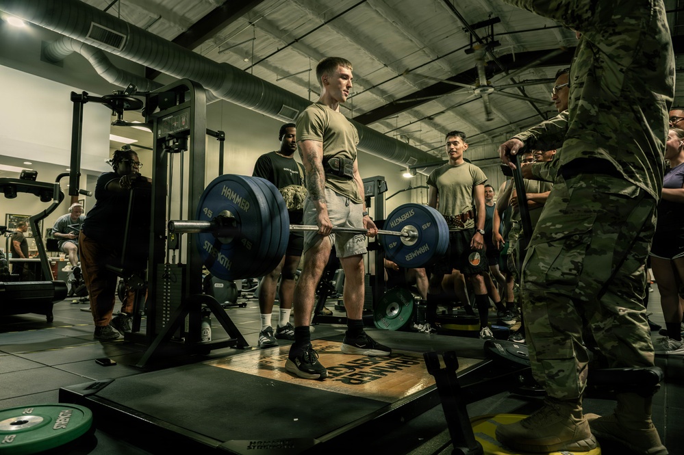 A military fitness setting where a soldier in an olive-green t-shirt and shorts is performing a deadlift with Hammer Strength 45-lb plates on a barbell. The lifter is wearing a weightlifting belt and stands on a deadlift platform, surrounded by observers in both PT gear and full camouflage uniforms. The scene appears to be a structured strength assessment or competition event within a military gymnasium.