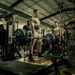 A military fitness setting where a soldier in an olive-green t-shirt and shorts is performing a deadlift with Hammer Strength 45-lb plates on a barbell. The lifter is wearing a weightlifting belt and stands on a deadlift platform, surrounded by observers in both PT gear and full camouflage uniforms. The scene appears to be a structured strength assessment or competition event within a military gymnasium.