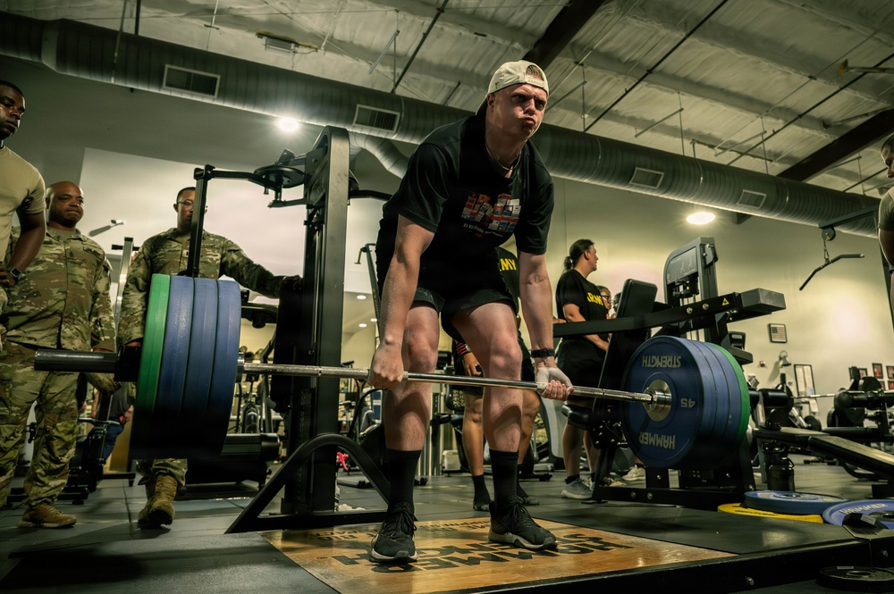 A person in a black t-shirt and backwards cap is performing a deadlift with a heavily loaded barbell in a military gym facility. Several individuals in U.S. Army camouflage uniforms (ACUs) are observing in the background. The setting appears to be an official military fitness center, likely during a strength competition or fitness assessment.