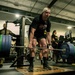 A person in a black t-shirt and backwards cap is performing a deadlift with a heavily loaded barbell in a military gym facility. Several individuals in U.S. Army camouflage uniforms (ACUs) are observing in the background. The setting appears to be an official military fitness center, likely during a strength competition or fitness assessment.