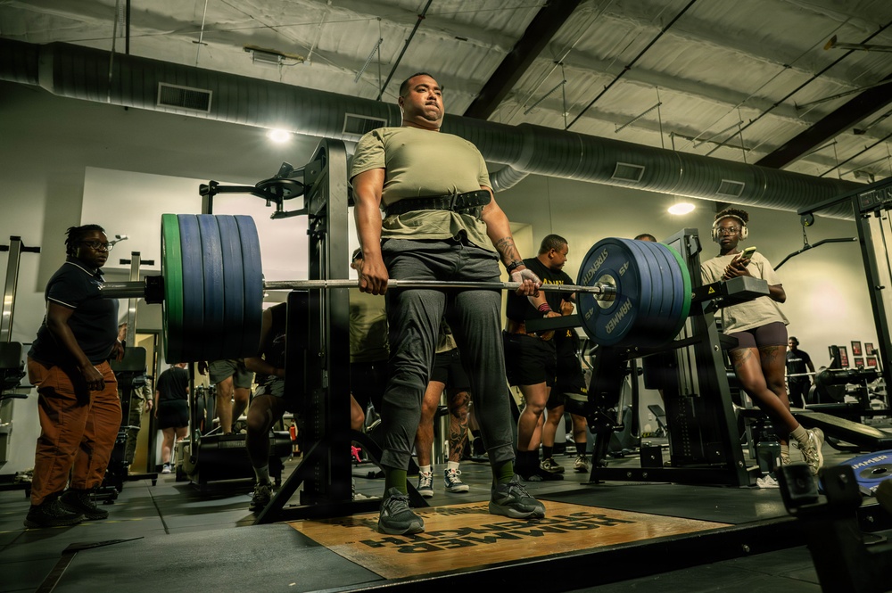 A powerlifter is performing a deadlift in a well-equipped gym, lifting a heavily loaded barbell with large bumper plates. Several other individuals are present in the background, some observing and others engaged in their own workouts. The lifter is wearing a weightlifting belt and appears to be mid-lift on a dedicated lifting platform.