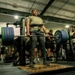 A powerlifter is performing a deadlift in a well-equipped gym, lifting a heavily loaded barbell with large bumper plates. Several other individuals are present in the background, some observing and others engaged in their own workouts. The lifter is wearing a weightlifting belt and appears to be mid-lift on a dedicated lifting platform.