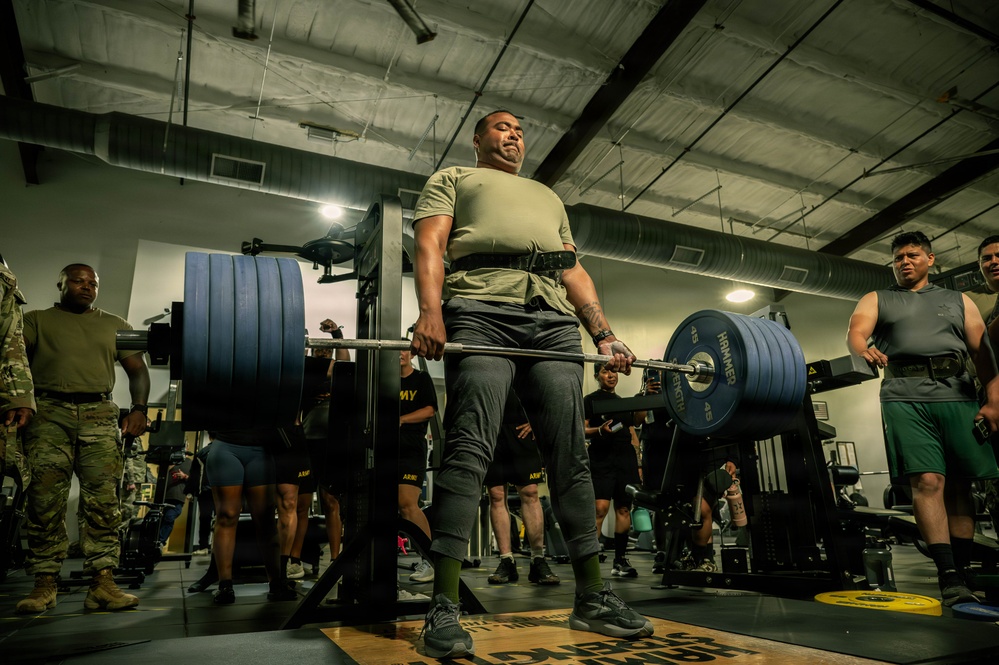 A U.S. Army soldier is performing a deadlift with a heavily loaded barbell in a military gym facility. The lifter is wearing a weightlifting belt and Army-branded athletic wear, surrounded by fellow soldiers observing the lift. The setting appears to be an organized strength training event or physical fitness assessment.