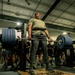 A U.S. Army soldier is performing a deadlift with a heavily loaded barbell in a military gym facility. The lifter is wearing a weightlifting belt and Army-branded athletic wear, surrounded by fellow soldiers observing the lift. The setting appears to be an organized strength training event or physical fitness assessment.