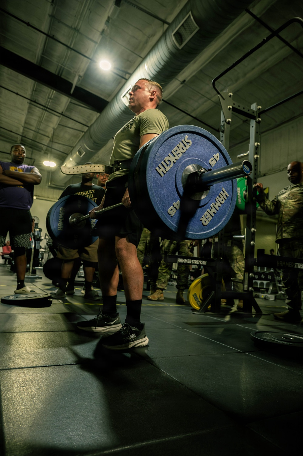 A military service member in an olive/tan PT shirt and black shorts performs a deadlift in a gym facility, holding a barbell loaded with Hammer Strength 45-lb bumper plates. Several spectators and other participants are visible in the background, some in military camouflage uniforms, suggesting a competitive or organized fitness event. The setting appears to be a military base gymnasium equipped with Hammer Strength branded equipment.