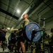A military service member in an olive/tan PT shirt and black shorts performs a deadlift in a gym facility, holding a barbell loaded with Hammer Strength 45-lb bumper plates. Several spectators and other participants are visible in the background, some in military camouflage uniforms, suggesting a competitive or organized fitness event. The setting appears to be a military base gymnasium equipped with Hammer Strength branded equipment.