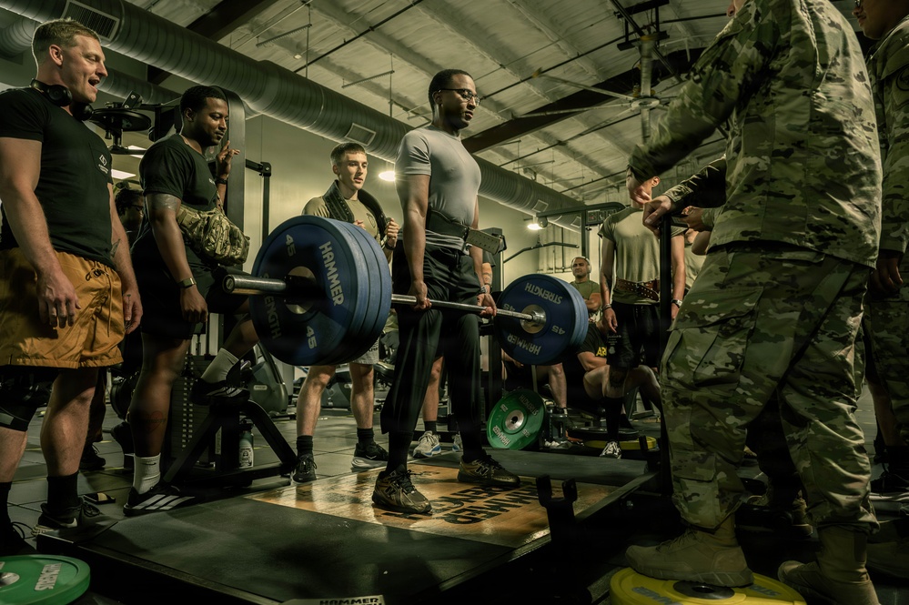 A military fitness setting where a soldier in a gray t-shirt is performing a deadlift with a heavily loaded barbell on a lifting platform. Multiple military personnel in various uniforms and PT gear are gathered around observing, suggesting a formal fitness test or competition event. The setting appears to be a well-equipped military gymnasium with industrial-style ceiling and ductwork.