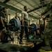 A military fitness setting where a soldier in a gray t-shirt is performing a deadlift with a heavily loaded barbell on a lifting platform. Multiple military personnel in various uniforms and PT gear are gathered around observing, suggesting a formal fitness test or competition event. The setting appears to be a well-equipped military gymnasium with industrial-style ceiling and ductwork.
