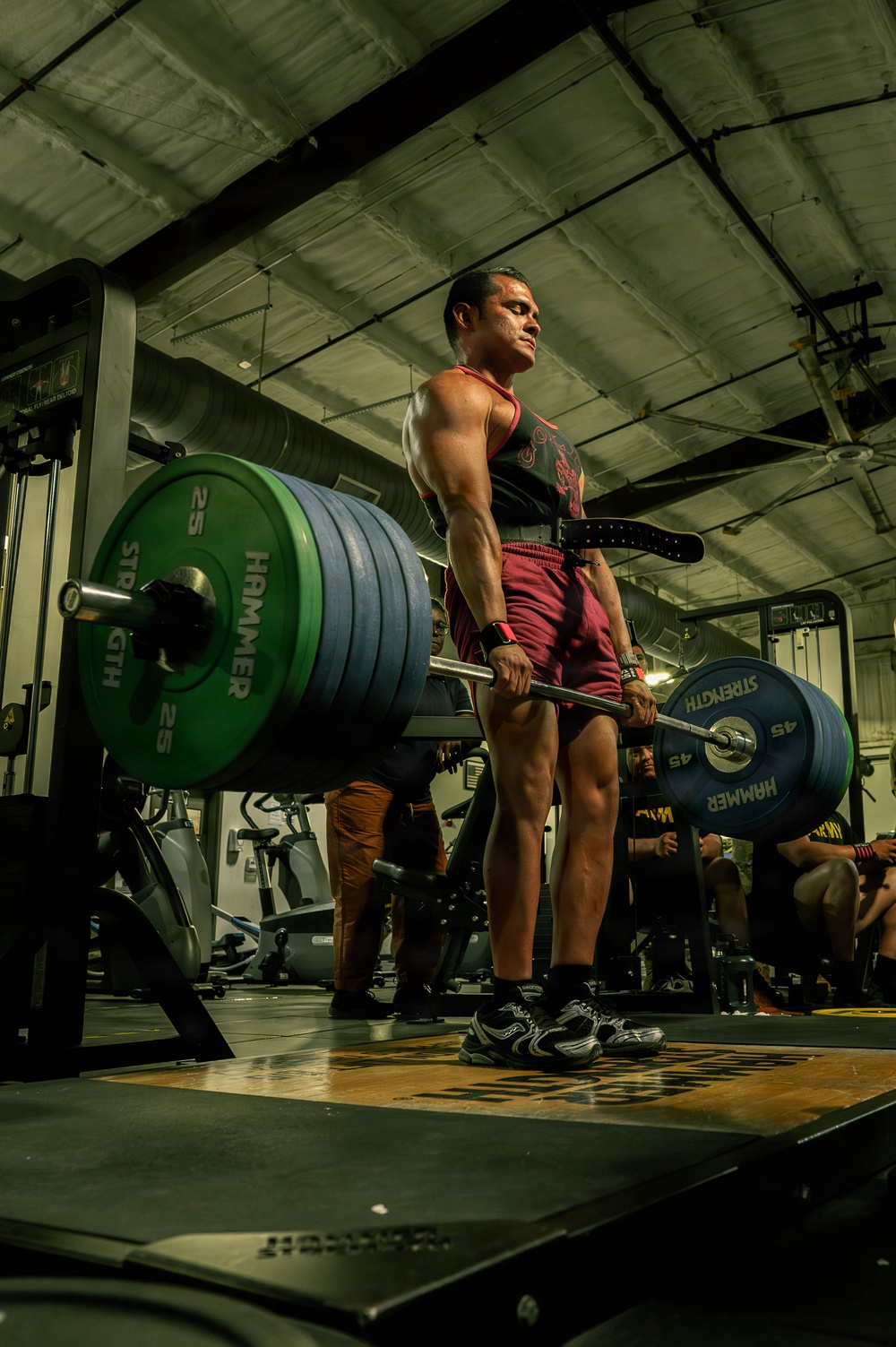 A muscular athlete performs a deadlift in an industrial-style gym, holding a heavily loaded barbell at hip level. The lifter wears a weightlifting belt, red tank top, maroon shorts, and athletic shoes, with a smartwatch visible on the wrist. Several onlookers and gym equipment are visible in the background.