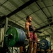 A muscular athlete performs a deadlift in an industrial-style gym, holding a heavily loaded barbell at hip level. The lifter wears a weightlifting belt, red tank top, maroon shorts, and athletic shoes, with a smartwatch visible on the wrist. Several onlookers and gym equipment are visible in the background.