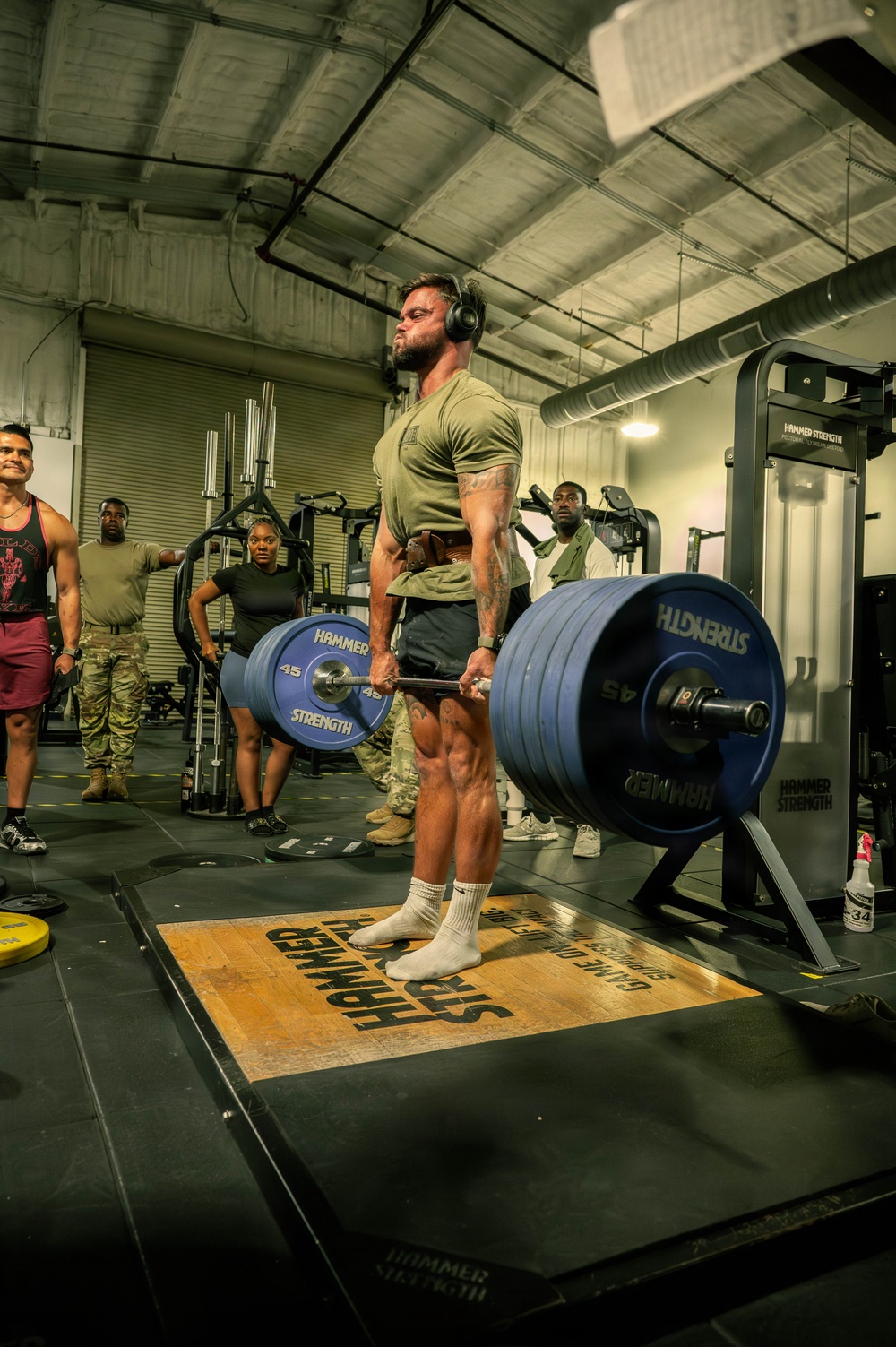 A muscular man wearing a green military-style t-shirt and headphones performs a deadlift on a Hammer Strength platform in an indoor gym facility. He is lifting a heavily loaded barbell with multiple 45 lb Hammer Strength bumper plates on each side. Several onlookers, some in military camouflage uniforms, observe the lift in the background.