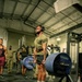 A muscular man wearing a green military-style t-shirt and headphones performs a deadlift on a Hammer Strength platform in an indoor gym facility. He is lifting a heavily loaded barbell with multiple 45 lb Hammer Strength bumper plates on each side. Several onlookers, some in military camouflage uniforms, observe the lift in the background.