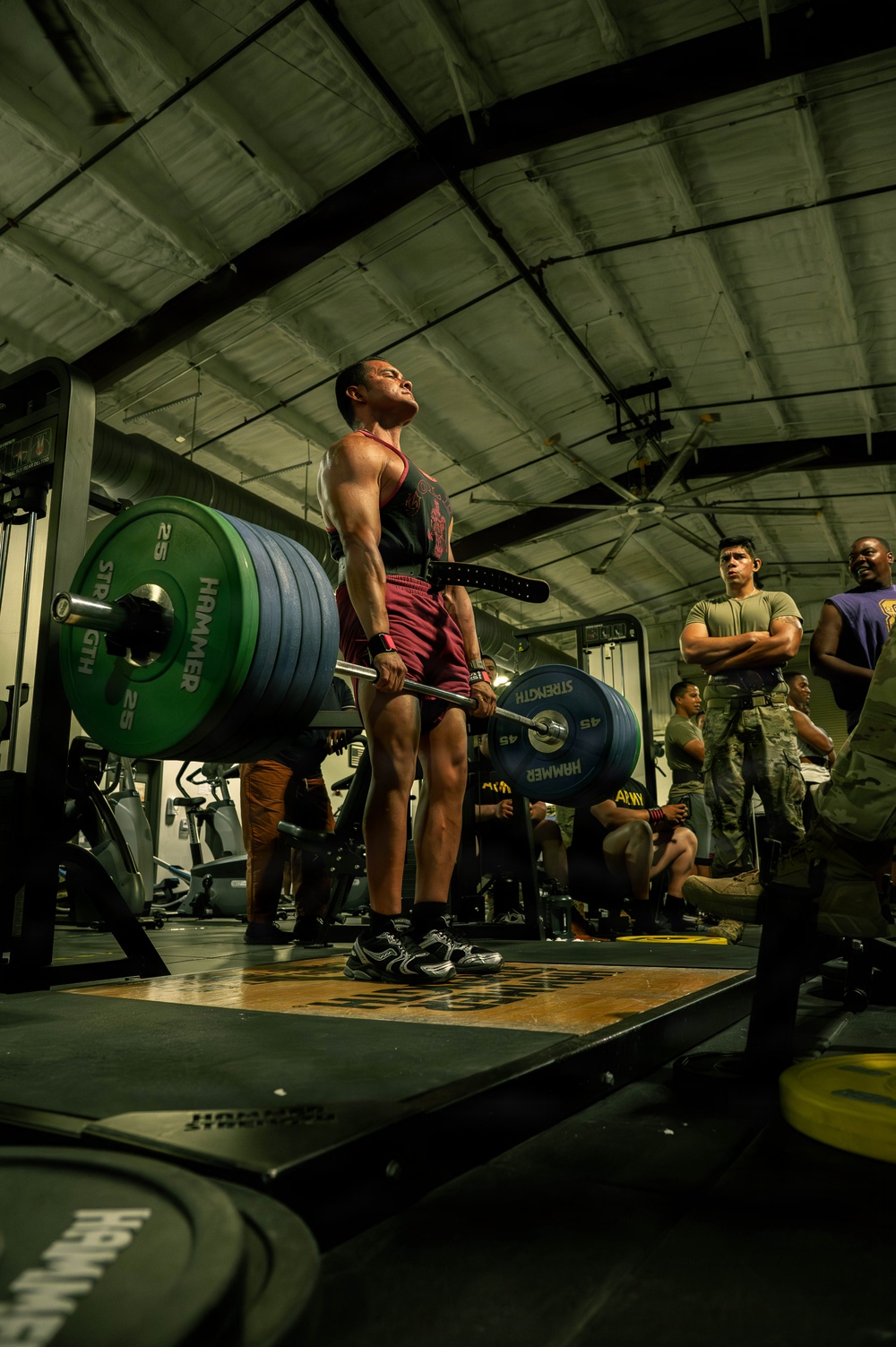 A muscular athlete performs a deadlift during what appears to be a military fitness competition, holding a heavily loaded barbell with multiple Hammer Strength weight plates. The lifter is positioned on a wooden lifting platform surrounded by spectators in military attire. The setting is an indoor military gymnasium with industrial ceiling and cardio equipment visible in the background.