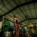 A muscular athlete performs a deadlift during what appears to be a military fitness competition, holding a heavily loaded barbell with multiple Hammer Strength weight plates. The lifter is positioned on a wooden lifting platform surrounded by spectators in military attire. The setting is an indoor military gymnasium with industrial ceiling and cardio equipment visible in the background.