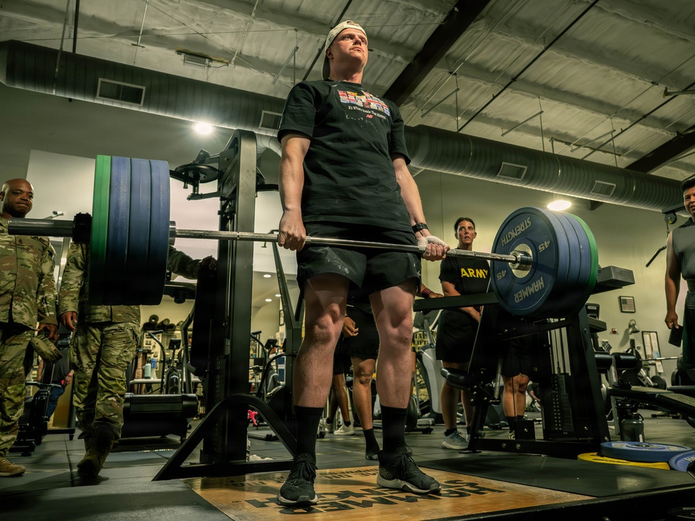 A person in military-affiliated athletic wear is performing a deadlift in a gym facility, holding a heavily loaded barbell with multiple 45-pound plates. Several observers, some in military camouflage uniforms, are watching from the sides. The setting appears to be a military fitness center based on the 'ARMY' branding visible on equipment.