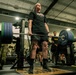 A person in military-affiliated athletic wear is performing a deadlift in a gym facility, holding a heavily loaded barbell with multiple 45-pound plates. Several observers, some in military camouflage uniforms, are watching from the sides. The setting appears to be a military fitness center based on the 'ARMY' branding visible on equipment.