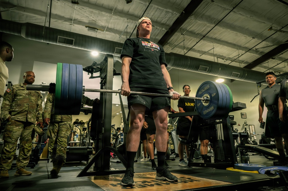 A soldier in civilian workout attire performs a deadlift with a heavily loaded barbell in a military fitness facility. Several observers, including personnel in Army Combat Uniforms (ACU/OCP pattern), watch from the sides. The setting appears to be an official Army gym, evidenced by 'ARMY HAMMER' branded weight plates and military insignia.