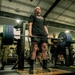 A soldier in civilian workout attire performs a deadlift with a heavily loaded barbell in a military fitness facility. Several observers, including personnel in Army Combat Uniforms (ACU/OCP pattern), watch from the sides. The setting appears to be an official Army gym, evidenced by 'ARMY HAMMER' branded weight plates and military insignia.