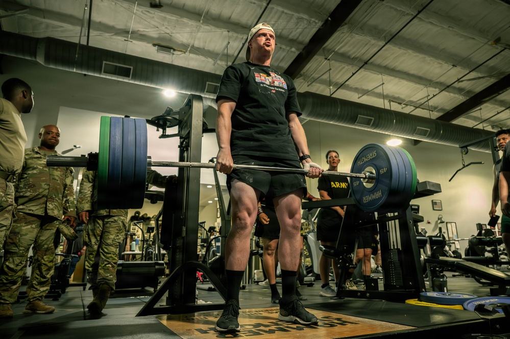 A soldier in civilian workout attire performs a deadlift with a heavily loaded barbell in a military gym facility. Several onlookers in U.S. Army combat uniforms (ACUs) observe the lift, suggesting a formal fitness event or competition. The barbell is branded with 'ARMY' and 'HAMMER STRENGTH' markings, and weight plates are loaded on both sides.