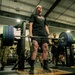 A soldier in civilian workout attire performs a deadlift with a heavily loaded barbell in a military gym facility. Several onlookers in U.S. Army combat uniforms (ACUs) observe the lift, suggesting a formal fitness event or competition. The barbell is branded with 'ARMY' and 'HAMMER STRENGTH' markings, and weight plates are loaded on both sides.