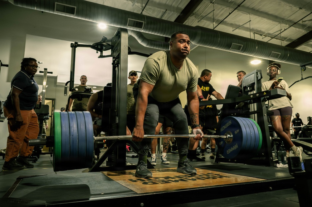 A muscular individual in a military-style olive green t-shirt is performing a deadlift in a gym setting, gripping a heavily loaded barbell with large green and blue weight plates. The setting appears to be a military fitness facility, evidenced by 'ARMY' branded clothing worn by several bystanders. Multiple observers are watching the lift in what appears to be either a training session or informal competition.