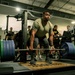 A muscular individual in a military-style olive green t-shirt is performing a deadlift in a gym setting, gripping a heavily loaded barbell with large green and blue weight plates. The setting appears to be a military fitness facility, evidenced by 'ARMY' branded clothing worn by several bystanders. Multiple observers are watching the lift in what appears to be either a training session or informal competition.