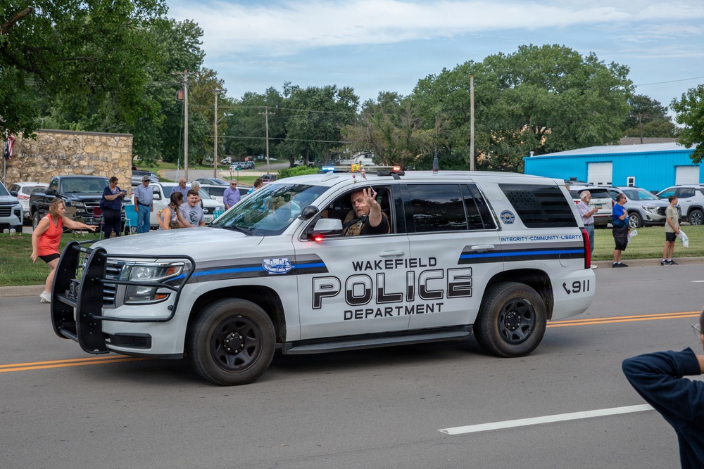 A Wakefield Police Department SUV patrol vehicle is participating in what appears to be a community parade or public event. An officer inside the vehicle is waving to bystanders lined along the street. The scene depicts a positive community-police interaction in a small-town setting.