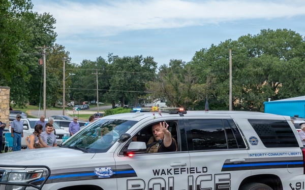 A Wakefield Police Department SUV patrol vehicle is participating in what appears to be a community parade or public event. An officer inside the vehicle is waving to bystanders lined along the street. The scene depicts a positive community-police interaction in a small-town setting.