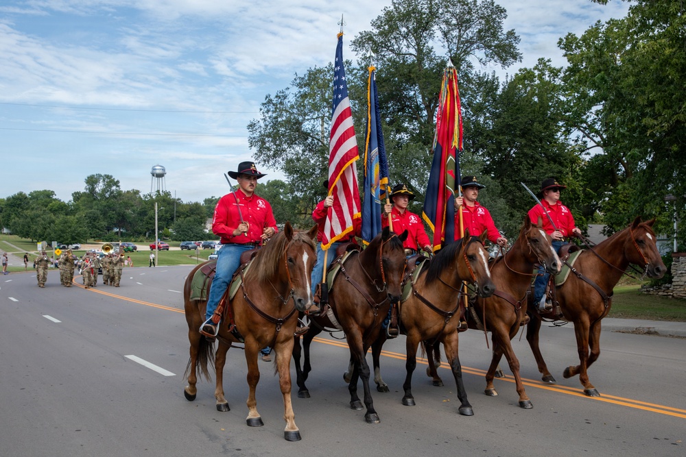 A military or ceremonial color guard on horseback participates in a parade, carrying the United States flag and additional military unit flags. The riders are dressed in matching red shirts and black cowboy hats, leading a procession down a tree-lined road. A military marching band in camouflage uniforms is visible in the background.