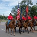A military or ceremonial color guard on horseback participates in a parade, carrying the United States flag and additional military unit flags. The riders are dressed in matching red shirts and black cowboy hats, leading a procession down a tree-lined road. A military marching band in camouflage uniforms is visible in the background.