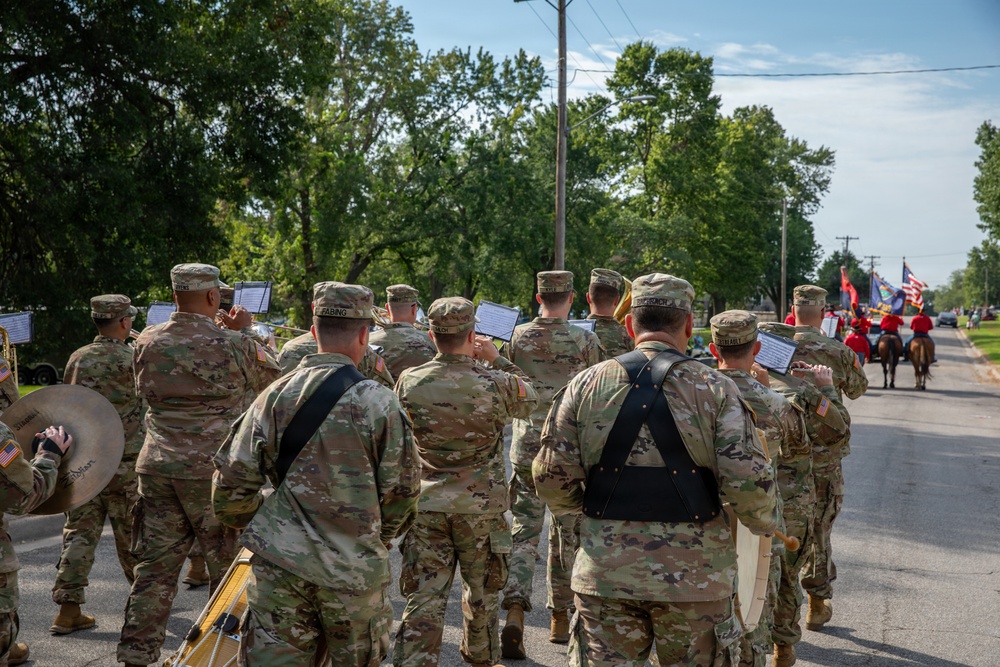 A U.S. Army military marching band is participating in an outdoor parade along a tree-lined street. The soldiers are dressed in OCP (Operational Camouflage Pattern) uniforms and are carrying instruments and music stands while marching. Ahead of them, riders on horseback and color guard members carrying flags are visible further down the parade route.