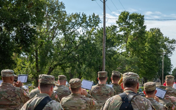 A U.S. Army military marching band is participating in an outdoor parade along a tree-lined street. The soldiers are dressed in OCP (Operational Camouflage Pattern) uniforms and are carrying instruments and music stands while marching. Ahead of them, riders on horseback and color guard members carrying flags are visible further down the parade route.