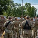 A U.S. Army military marching band is participating in an outdoor parade along a tree-lined street. The soldiers are dressed in OCP (Operational Camouflage Pattern) uniforms and are carrying instruments and music stands while marching. Ahead of them, riders on horseback and color guard members carrying flags are visible further down the parade route.