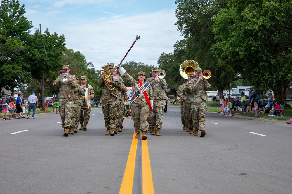 A military marching band in camouflage uniforms performs during a street parade on a sunny day. The band includes brass instrument players, a drum major leading with a baton, and percussion players marching down a two-lane road lined with spectators. The setting appears to be a small-town community parade with families watching from the sidewalks.
