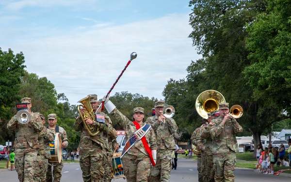 A military marching band in camouflage uniforms performs during a street parade on a sunny day. The band includes brass instrument players, a drum major leading with a baton, and percussion players marching down a two-lane road lined with spectators. The setting appears to be a small-town community parade with families watching from the sidewalks.