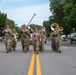 A military marching band in camouflage uniforms performs during a street parade on a sunny day. The band includes brass instrument players, a drum major leading with a baton, and percussion players marching down a two-lane road lined with spectators. The setting appears to be a small-town community parade with families watching from the sidewalks.
