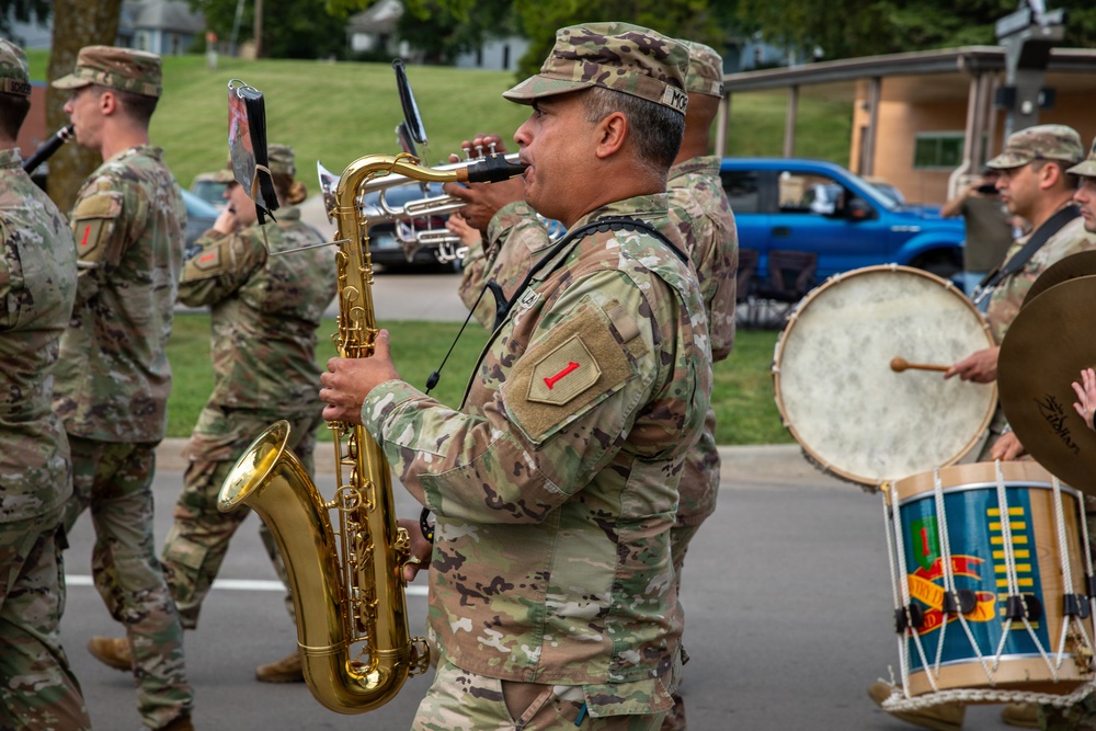 A U.S. Army military band is marching in a parade or ceremony, with soldiers in OCP (Operational Camouflage Pattern) uniforms playing various instruments. The central figure is playing a tenor saxophone, while others play trumpet and bass drum. The patch on the saxophone player's shoulder appears to be the 1st Infantry Division ('Big Red One') insignia.