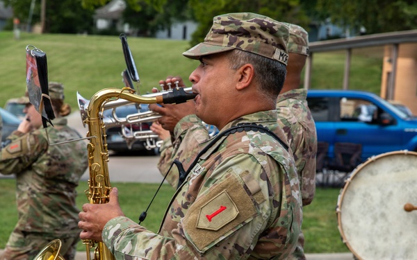 A U.S. Army military band is marching in a parade or ceremony, with soldiers in OCP (Operational Camouflage Pattern) uniforms playing various instruments. The central figure is playing a tenor saxophone, while others play trumpet and bass drum. The patch on the saxophone player's shoulder appears to be the 1st Infantry Division ('Big Red One') insignia.