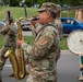 A U.S. Army military band is marching in a parade or ceremony, with soldiers in OCP (Operational Camouflage Pattern) uniforms playing various instruments. The central figure is playing a tenor saxophone, while others play trumpet and bass drum. The patch on the saxophone player's shoulder appears to be the 1st Infantry Division ('Big Red One') insignia.