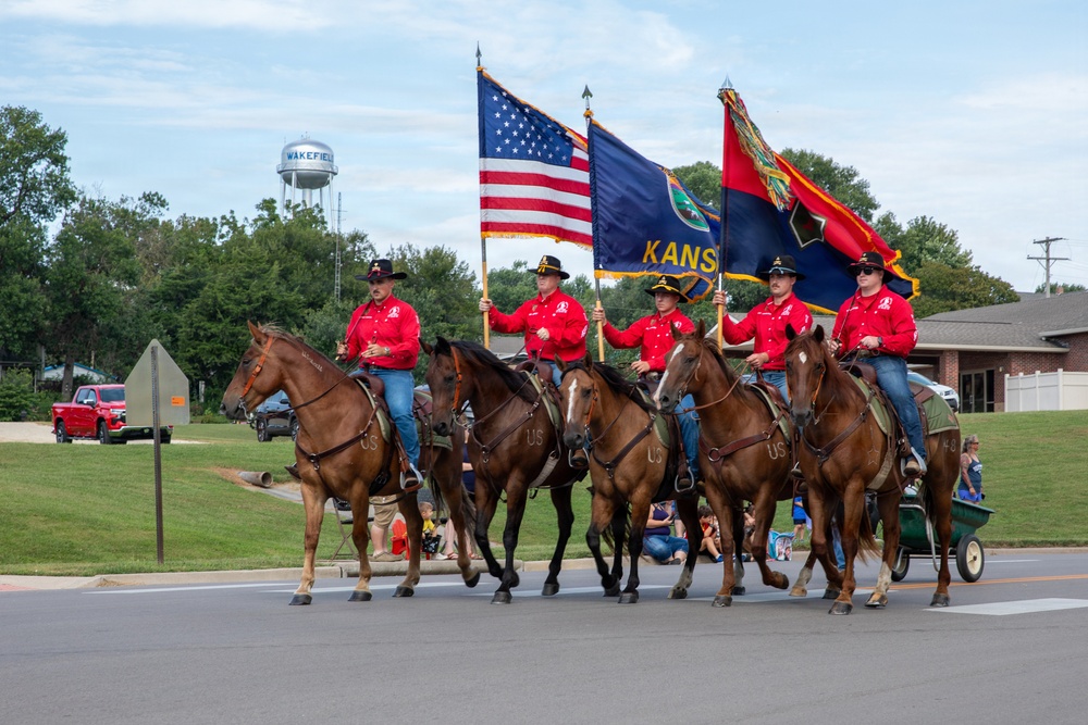 A military or ceremonial color guard on horseback participates in a parade in Wakefield, Kansas. Five riders dressed in matching red shirts and black cowboy hats carry the American flag, the Kansas state flag, and what appears to be a military unit flag. The water tower in the background clearly identifies the location as Wakefield, and spectators line the parade route.