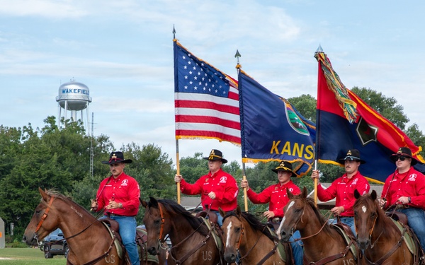 A military or ceremonial color guard on horseback participates in a parade in Wakefield, Kansas. Five riders dressed in matching red shirts and black cowboy hats carry the American flag, the Kansas state flag, and what appears to be a military unit flag. The water tower in the background clearly identifies the location as Wakefield, and spectators line the parade route.