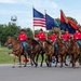 A military or ceremonial color guard on horseback participates in a parade in Wakefield, Kansas. Five riders dressed in matching red shirts and black cowboy hats carry the American flag, the Kansas state flag, and what appears to be a military unit flag. The water tower in the background clearly identifies the location as Wakefield, and spectators line the parade route.