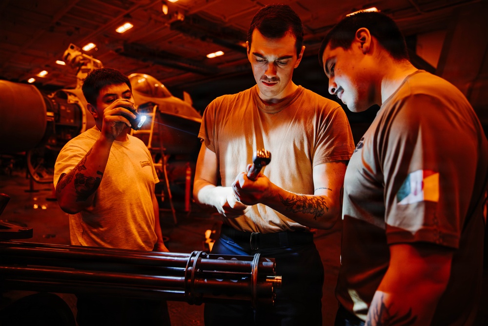Three military personnel are working in what appears to be a dimly lit hangar bay, likely aboard a naval vessel. They are examining a multi-barrel rotary cannon component (consistent with a Gatling-style weapon system) using flashlights. The scene is illuminated primarily by warm orange ambient lighting with aircraft visible in the background.