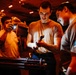 Three military personnel are working in what appears to be a dimly lit hangar bay, likely aboard a naval vessel. They are examining a multi-barrel rotary cannon component (consistent with a Gatling-style weapon system) using flashlights. The scene is illuminated primarily by warm orange ambient lighting with aircraft visible in the background.