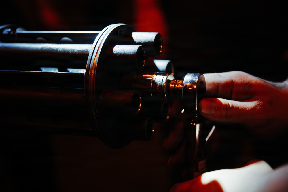 A close-up photograph shows a human hand touching or handling a multi-barrel rotary weapon, consistent with a Gatling-style gun or minigun configuration. The image is dramatically lit with dark shadows and warm red/orange ambient lighting highlighting the metallic barrels. Multiple cylindrical gun barrels are visible arranged in a circular cluster pattern.