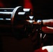 A close-up photograph shows a human hand touching or handling a multi-barrel rotary weapon, consistent with a Gatling-style gun or minigun configuration. The image is dramatically lit with dark shadows and warm red/orange ambient lighting highlighting the metallic barrels. Multiple cylindrical gun barrels are visible arranged in a circular cluster pattern.