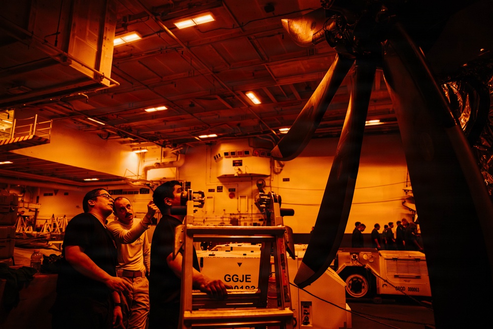 Three personnel are working in what appears to be a military vessel hangar bay, illuminated by orange/red lighting. They are inspecting or maintaining a large rotor assembly, likely from a helicopter, mounted on a support stand. The environment contains various equipment, crates, and machinery consistent with a naval aircraft maintenance facility.