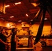 Three personnel are working in what appears to be a military vessel hangar bay, illuminated by orange/red lighting. They are inspecting or maintaining a large rotor assembly, likely from a helicopter, mounted on a support stand. The environment contains various equipment, crates, and machinery consistent with a naval aircraft maintenance facility.