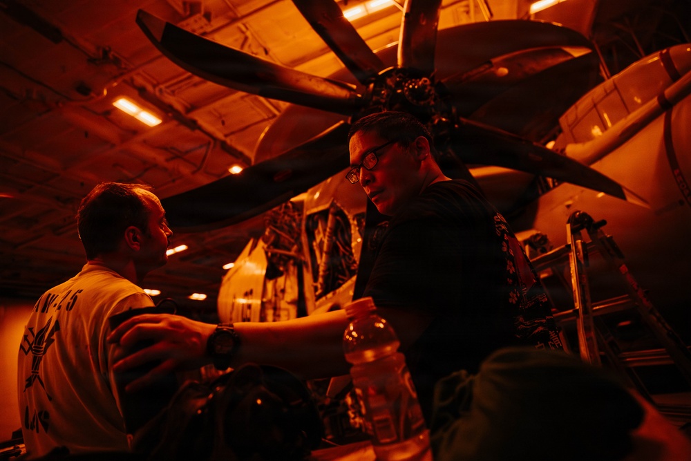 Two individuals are working in what appears to be an aircraft hangar bay, likely aboard a naval vessel, illuminated by orange/red lighting. A large multi-blade propeller aircraft is visible overhead, suggesting maintenance or inspection work is being conducted. The environment is enclosed with overhead lighting strips and mechanical infrastructure visible.