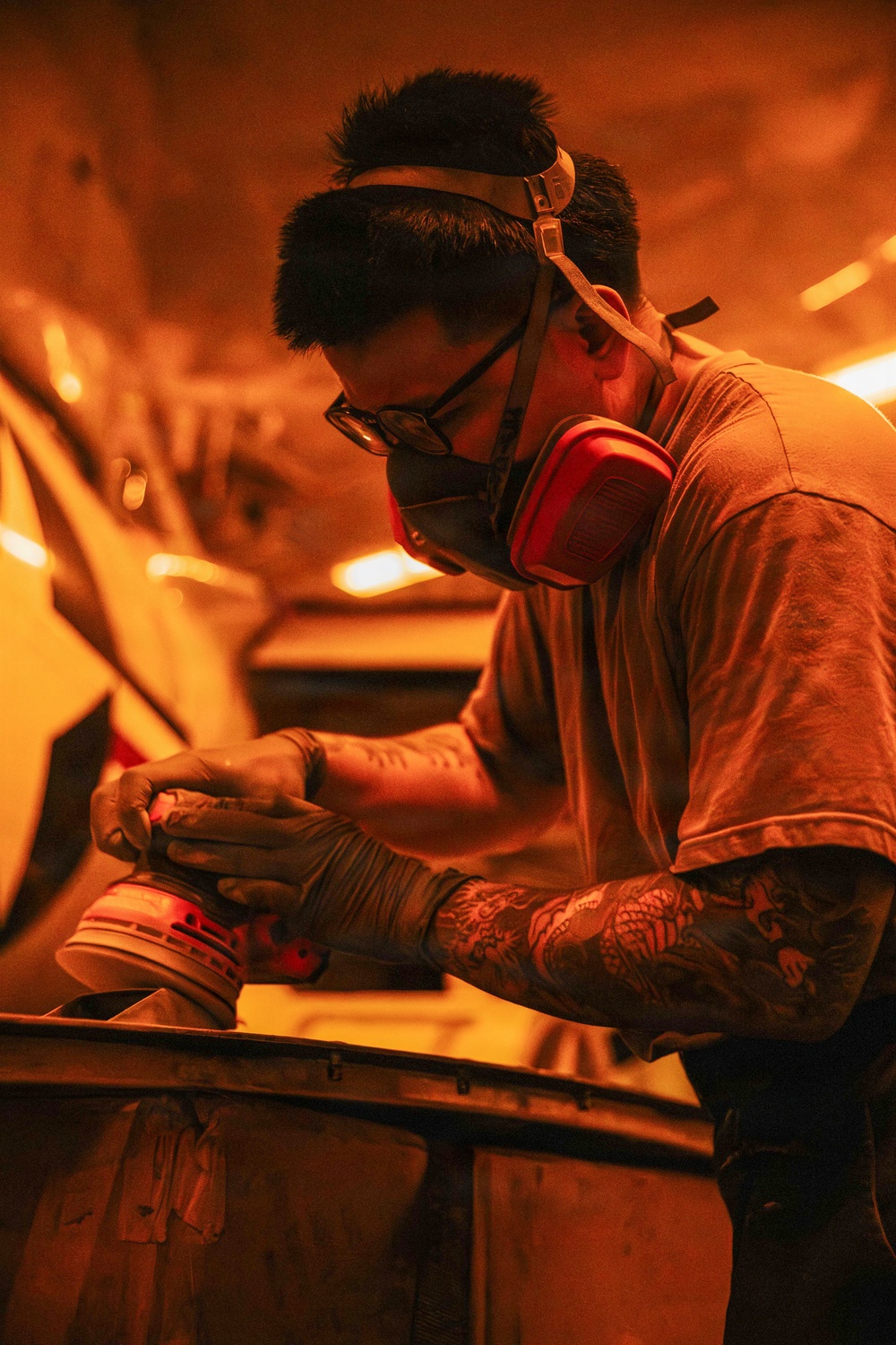 A tattooed male worker is using a power sander on what appears to be an automotive body panel in a workshop environment. He is wearing appropriate personal protective equipment including a dual-cartridge respirator mask, safety glasses, and work gloves. The scene is bathed in warm orange ambient lighting, likely from heat lamps used in auto body work.